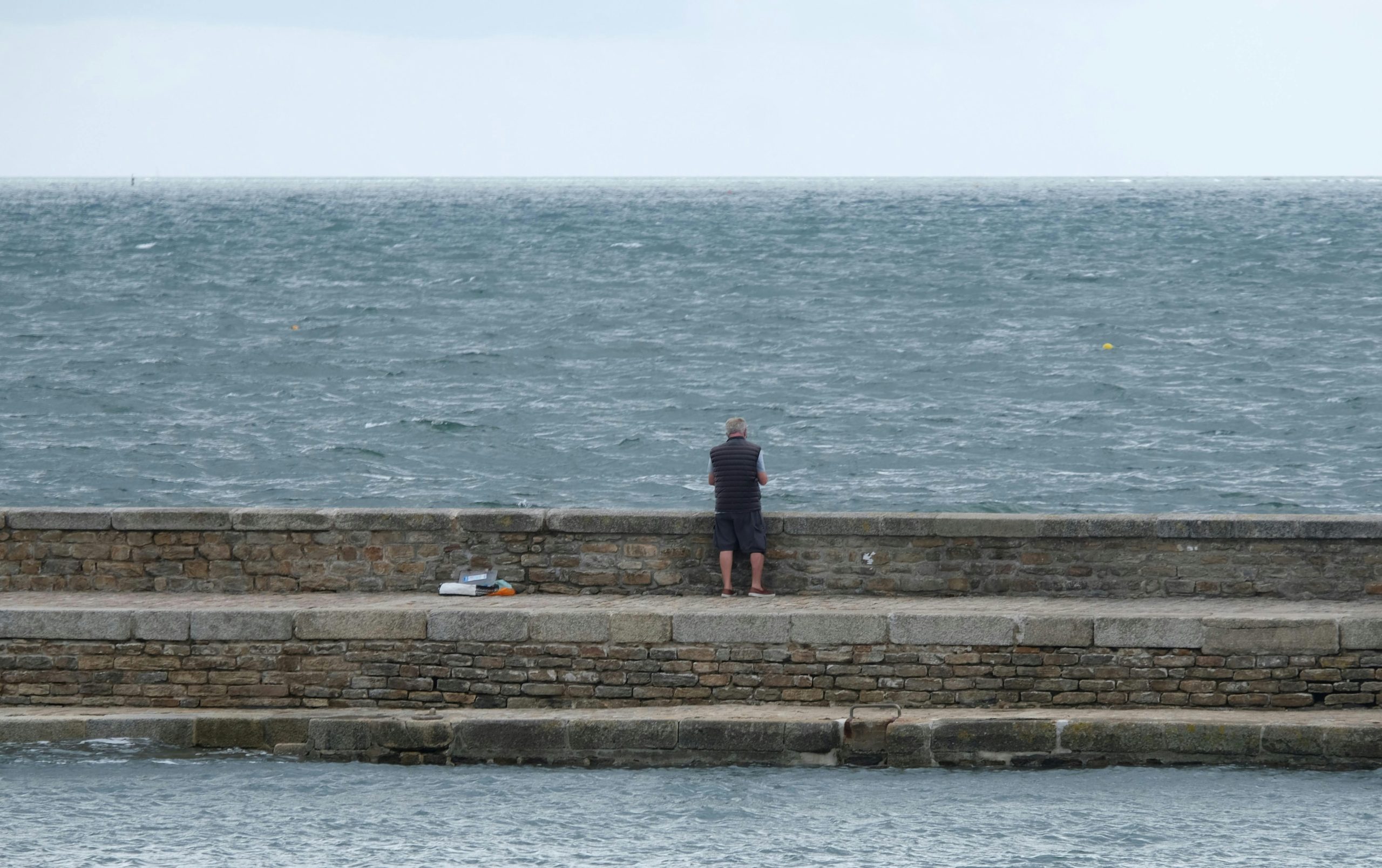 A solitary man stands on a stone pier overlooking the vast, tranquil sea.